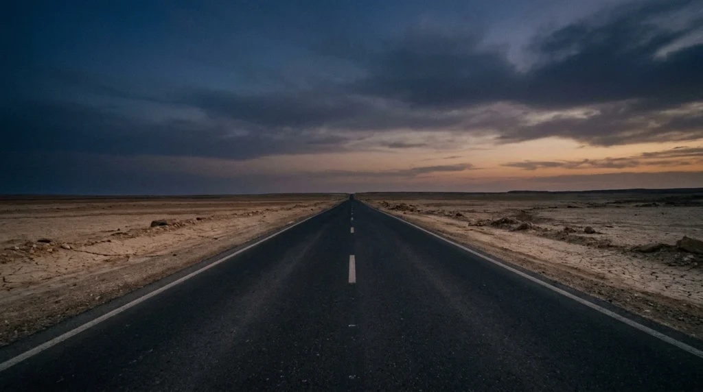 Empty unlit rural two-lane road stretching toward the horizon at dusk